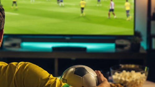 Pessoa assistindo a uma partida de futebol pela televisão em ambiente residencial, ilustrando a experiência de acompanhar jogos da Copa do Mundo.
