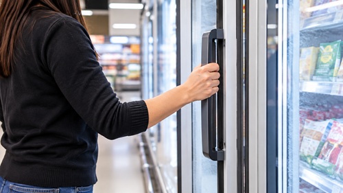 Mulher abre porta de refrigerador em supermercado, sem mostrar o rosto.